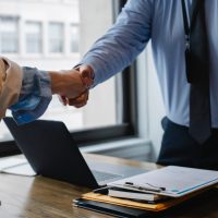 PPFD is a Proud Member of the Whitby Chamber of Commerce. News feature showing two professionals in business attire shaking hands over a desk with a laptop and documents, signifying a successful meeting or agreement.