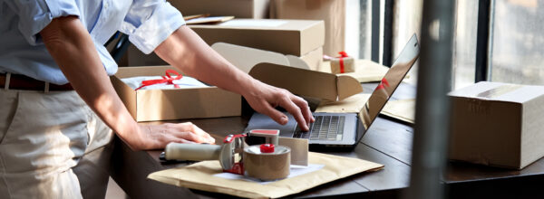 PPFD Delivery Guidelines for Distribution Centers in Canada. Product preparation with a person packing items into boxes at a workstation, with a laptop and packing tape at hand, indicative of a busy fulfillment and distribution environment.