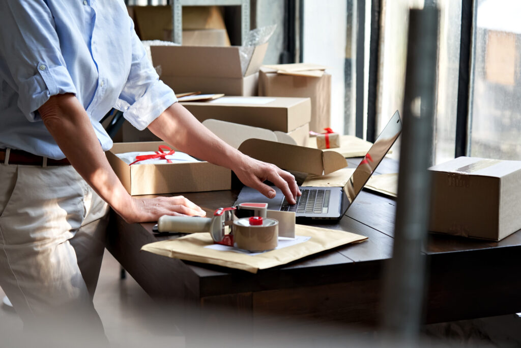 PPFD Delivery Guidelines for Distribution Centers in Canada. Product preparation with a person packing items into boxes at a workstation, with a laptop and packing tape at hand, indicative of a busy fulfillment and distribution environment.