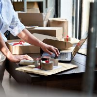 PPFD Delivery Guidelines for Distribution Centers in Canada. Product preparation with a person packing items into boxes at a workstation, with a laptop and packing tape at hand, indicative of a busy fulfillment and distribution environment.