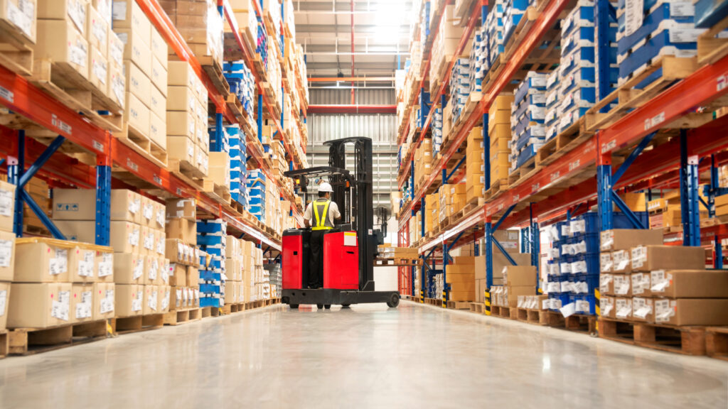 A wide-aisle warehouse interior with tall orange and blue shelving stacked with various boxes and a red forklift in the aisle operated by a worker in safety gear.