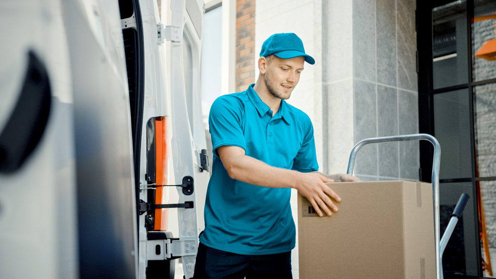 A delivery man in a blue uniform and cap is unloading a cardboard box from a van. He is smiling and handling the box carefully outside a building.