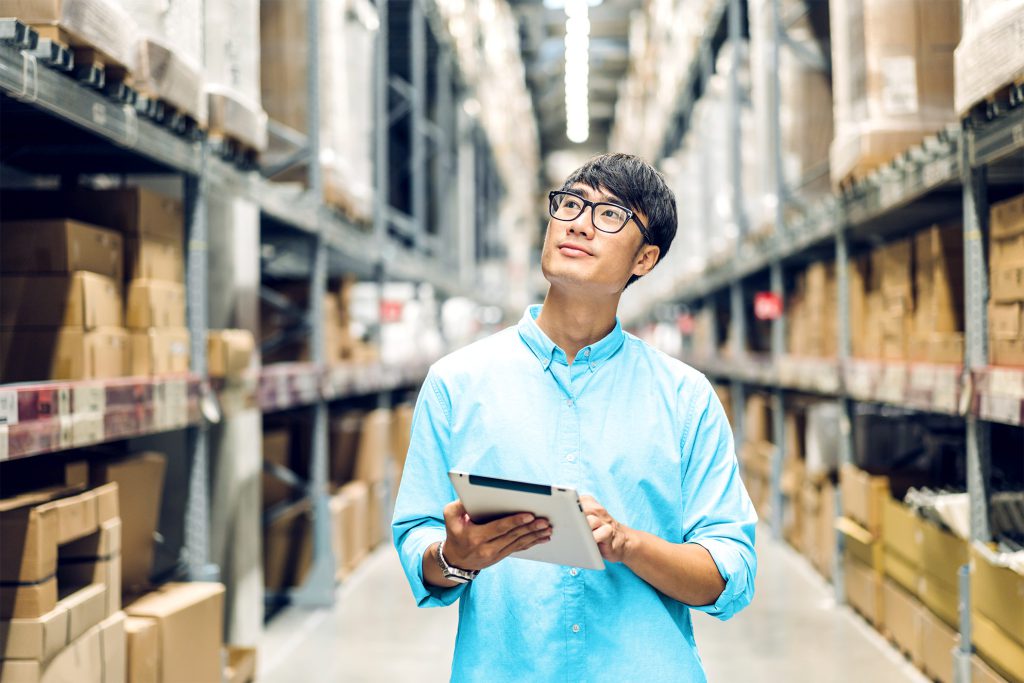 A man in a light blue shirt and glasses is inspecting the inventory in a large warehouse. He is holding a tablet and looking up at the shelves filled with boxes.